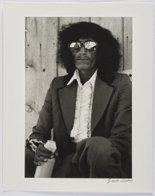 Black and white photograph of a man with shoulder-length hair, wearing reflective sunglasses, ruffled white shirt and suit with wide lapels, 