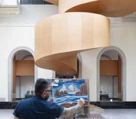 Man with black rimmed glasses seen from behind drawing an image of mountains on an easel.