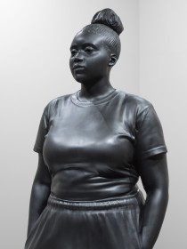 A bronze statue depicting an African American woman wearing a t-shirt, and sweatpants. Their hands are casually in their pockets and they are softly glancing to their right 