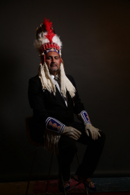A Photo of Adrian Stimson, seated. Adrian is wearing indigenous headwear, and indigenous gloves. Otherwise, he is wearing a black suit. 