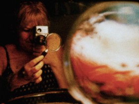 A still from Joyce Weiland's Water Sark. A female figure points a camera through a magnifying glass at a round, abstract subject. 