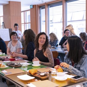 Group of people seated at a table with coffee and pastries, smiling in a bright room.