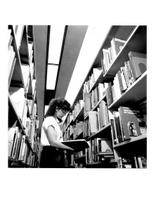 Black and white photo of young white woman with long hair and glasses reading a book between library shelves. From the 1980s