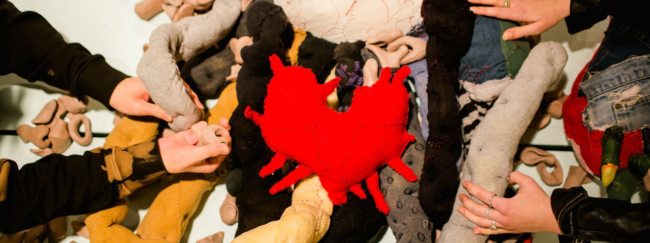 From above, a photo of two people touching a sprawling textile sculpture called Big Softie. They reach into the guts, which are soft and dimpled and made from stuffed knee socks and nylon stockings, and examine the Unidentified Remains that are scattered amongst them. At the centre lies Big Softie’s heart, a red patchwork soft sculpture with tendrils extending outward.