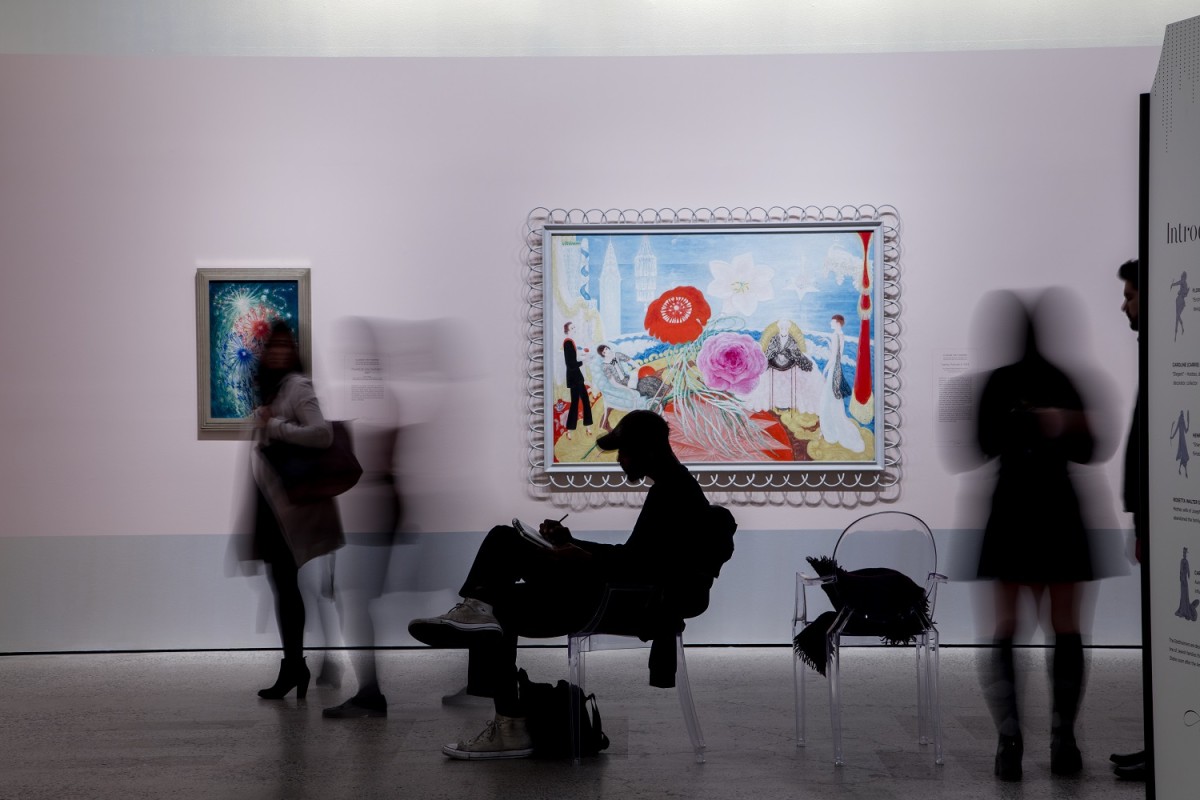 Man sits and sketches in front of Florine Stettheimer works, surrounded by other gallery visitors.