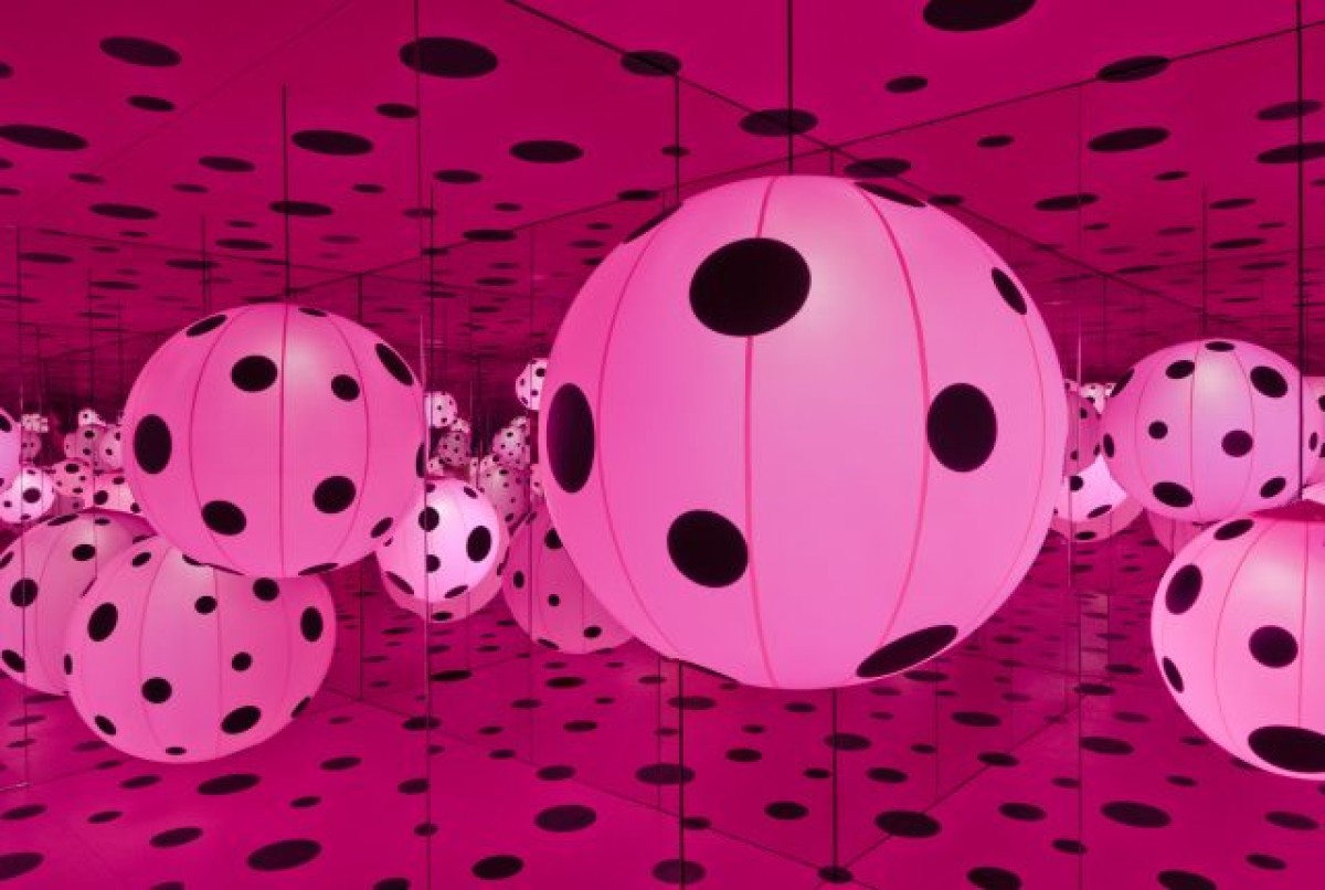 interior view of Yayoi Kusama's Dots Obsession Infinity Mirror Room, reflected endlessly in mirrors. Background is pink with black polka dots. 