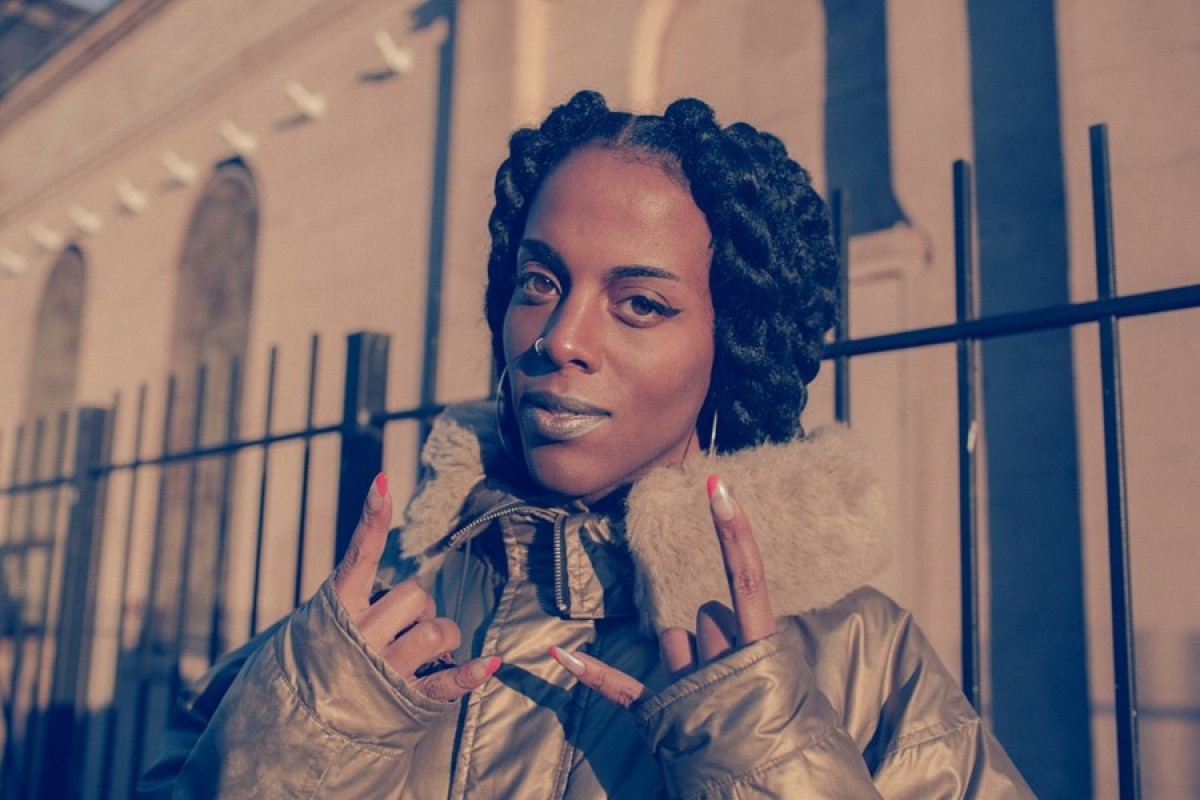 Artist Juliana Huxtable wears a brown winter coat and holds two nail-polished fingers up on each hand.