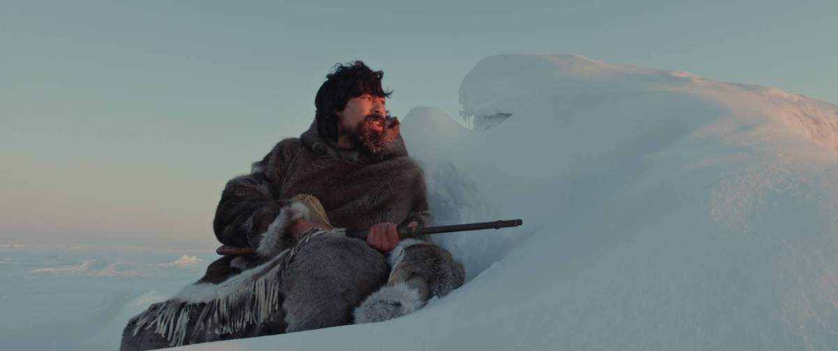 An Inuit man hides behind a bank of snow, with the light of the setting sun hitting his face.