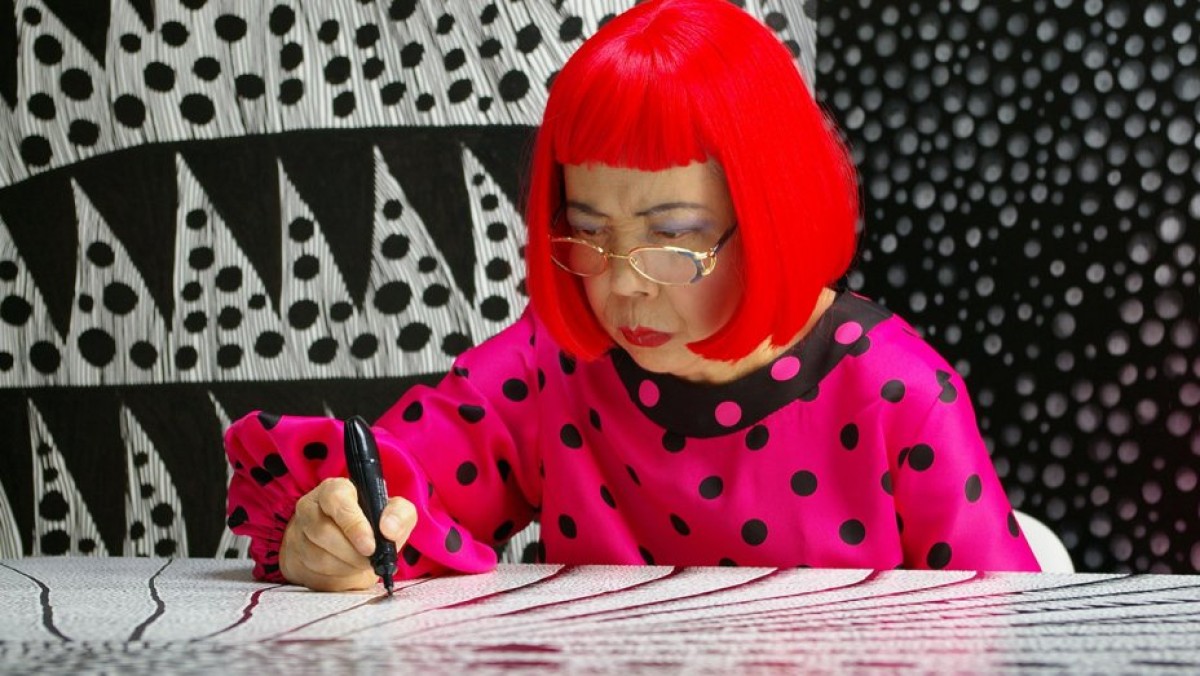 Artist Yayoi Kusama wears a bright red wig, glasses, red lips, and a pink and black polka-dotted dress and draws a black and white design against a black and white patterned backdrop.