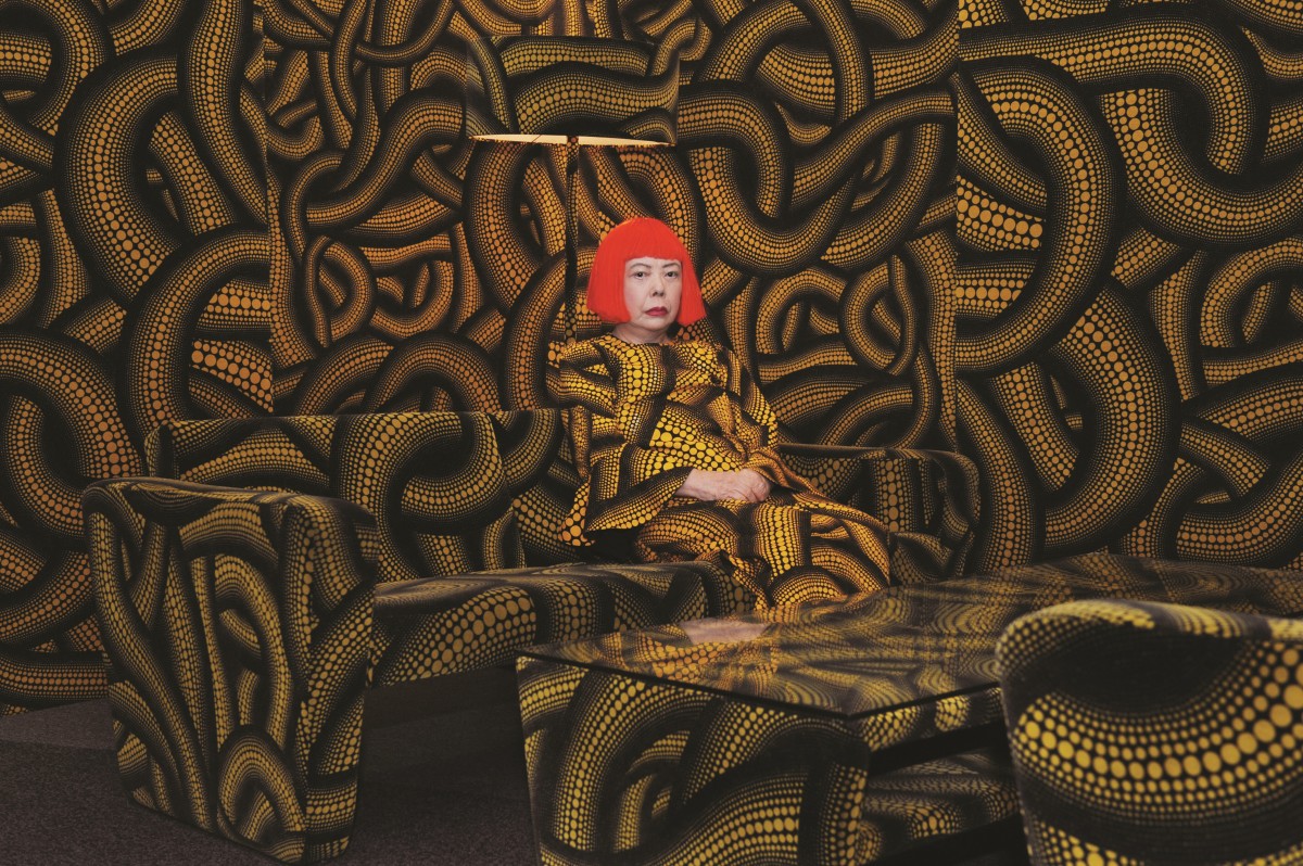 Yayoi Kusama wears a bright red wig while sitting in a room with the same black and yellow patter on the walls, furniture and clothing.
