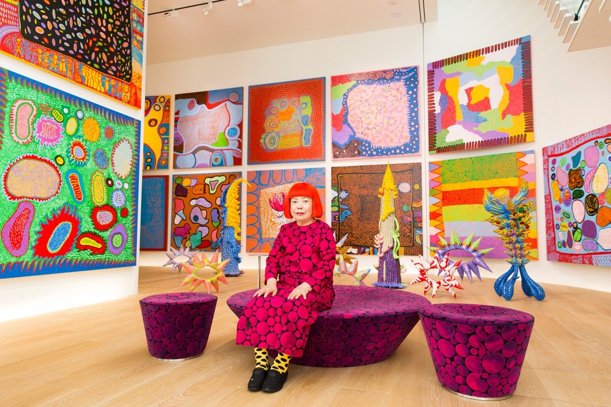Yayoi Kusama wears a red wig and purple polka dotted dress in a gallery surrounded by her sculptures and paintings.