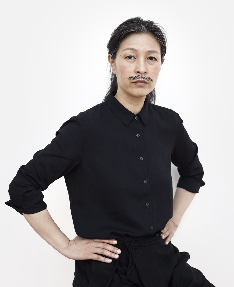 Artist Haeugue Yang poses with hands on hips, sporting a drawn on mustache 