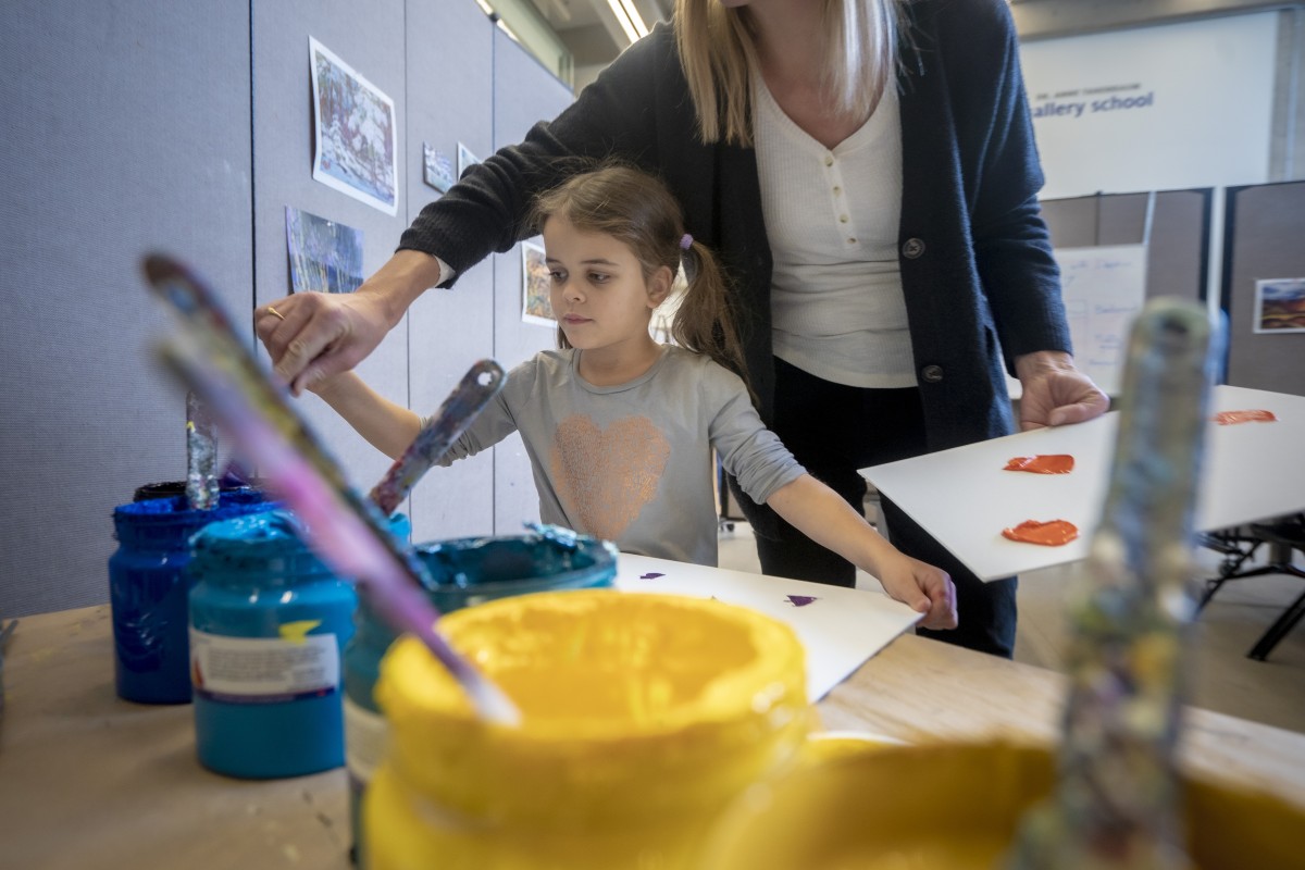 a girl scoops blue and yellow paint from large jars with the help of her mother. 