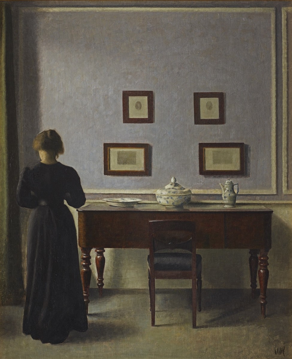 Painting of a woman with her back turned, standing in front of a table looking at a wall with four small paintings on it. 