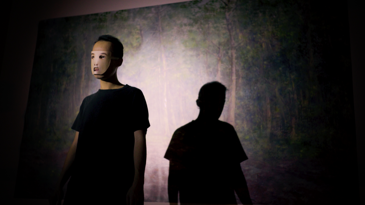 A man in a mask stands in a spotlight, in front of a painting of a forest. 