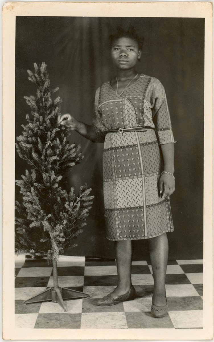 A woman wearing a patterned dress stands beside a small Christmas tree. She looks directly at the viewer.