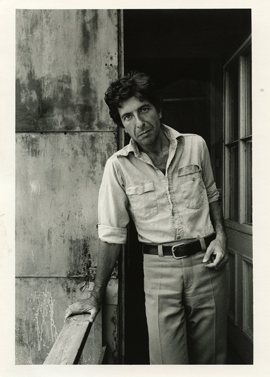 Black and white vertical photo of Leonard Cohen in 1973, wearing a casual collared shirt, standing with one hand on a banister, slightly leaning to his right and looking into the camera