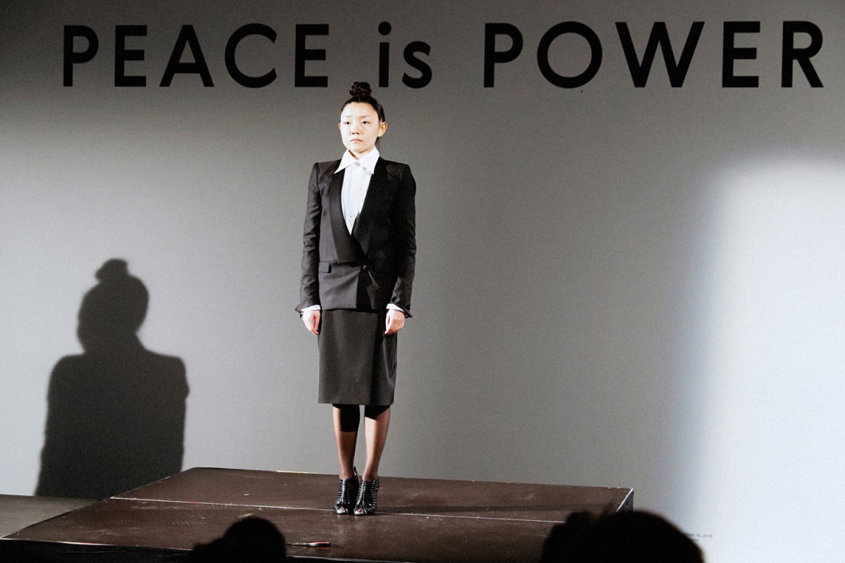 woman standing on stage dressed in a suit dress with "Peace is Power" written on a wall behind her