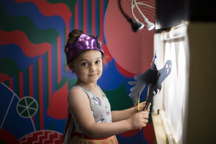 A young girl holds an owl shadow puppet behind a puppet theatre.