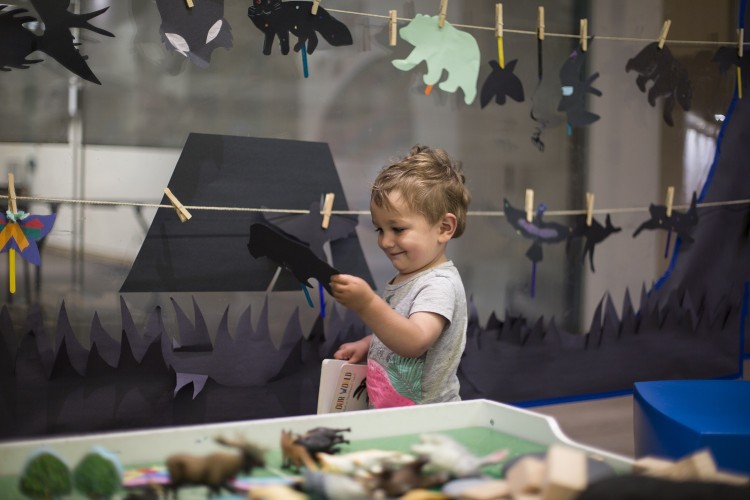 A young boy holds a bird shadow puppet as he walks in front of a laundry line of black paper puppets.