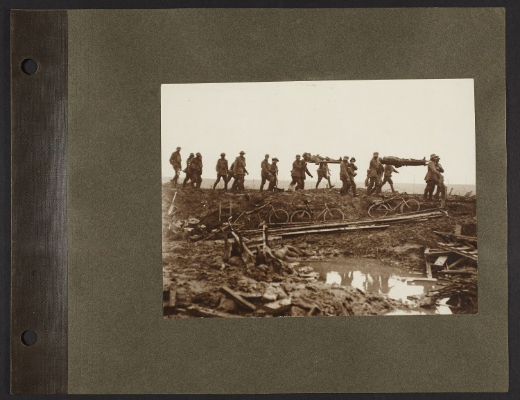 A black and white photograph of soldiers carrying two men in stretchers across a destroyed terrain.