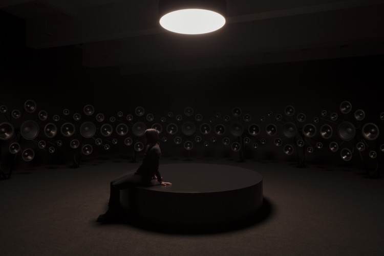 A person sits in a dark room alone, surrounded by speakers