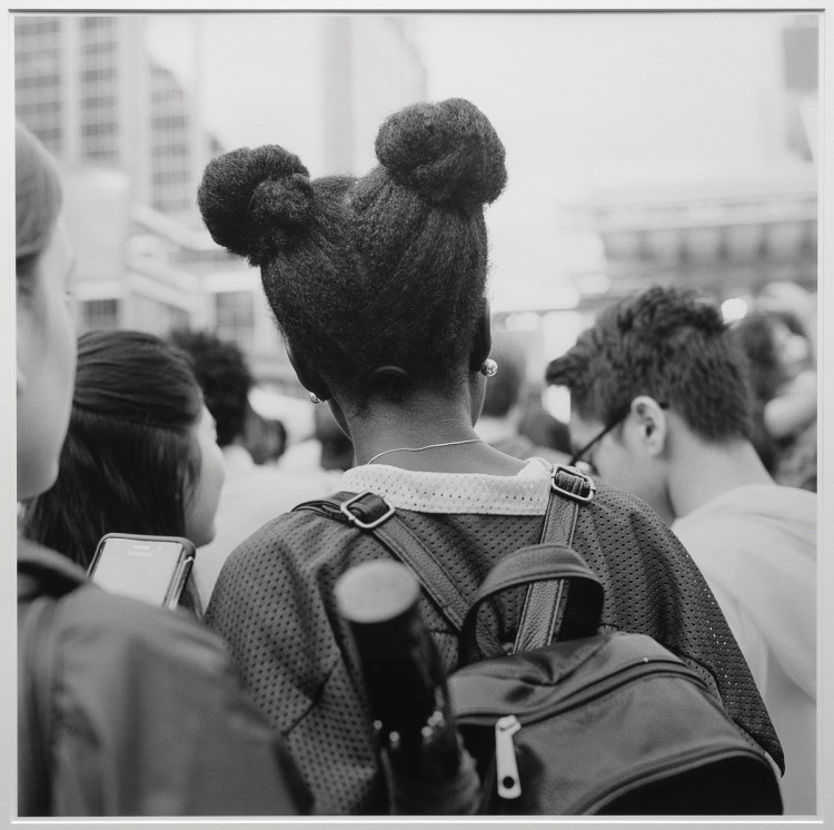 Black and white photo of the back of a young Black woman, wearing a backpack and hair in buns at each side of the head 
