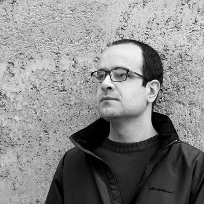 A picture of Morteza who stands looking away from the camera. He wears a black sweater, jacket and glasses. In the background, there is a wall with a rough texture.