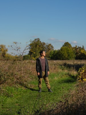 A figure wearing a brown shirt, grey sweater and khaki pants is standing in a field of grass, head turned to the right. Trees are lined up along the horizon against a clear, blue sky. 