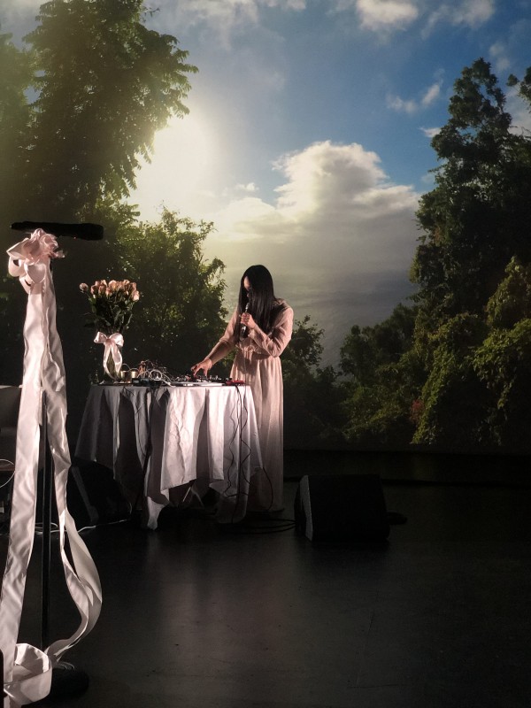 colour photo of performance featuring Miho Hatori in front of a green treed landscape, with a table draped in white with pink ribbons
