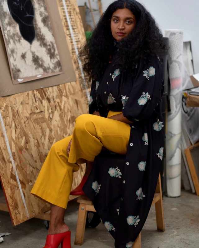 Artist Rajni Perera seated in her studio wearing a long black coat and yellow trousers