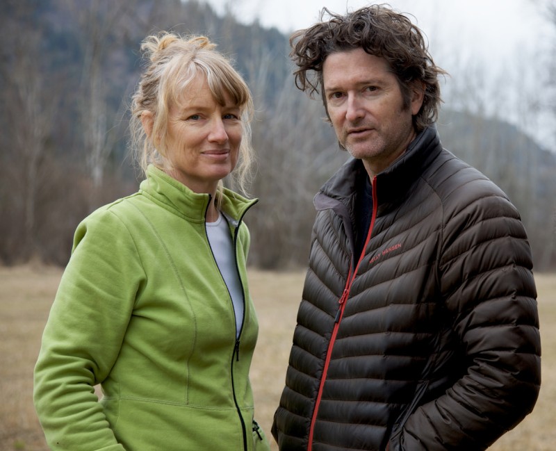 Artists Janet Cardiff in a green jacket and George Bures MIller in a black puffer jacket, standing in a field with a hill behind them. 