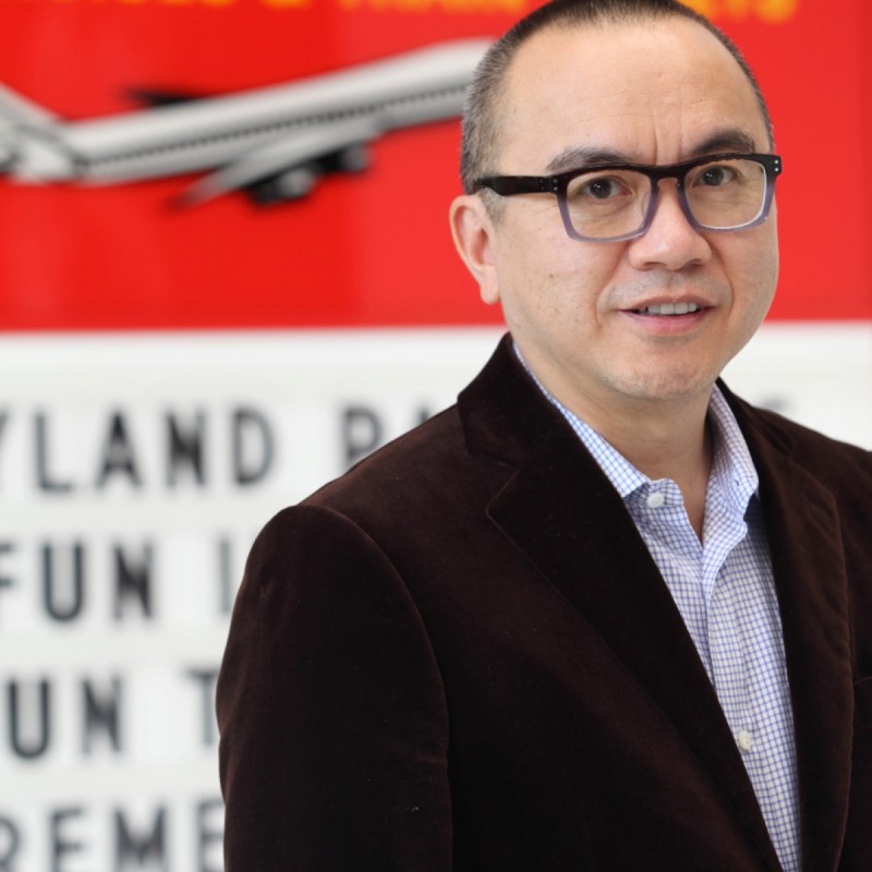 Artist Ken Lum in dark blazer, light blue shirt with red background with and airplane and black text