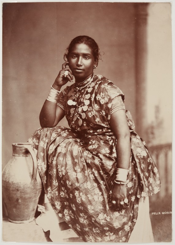 Photo of a Hindu woman taken in Trinidad, wearing a pattered dress, metal bracelets and layered necklaces, posing beside a tall clay vessel