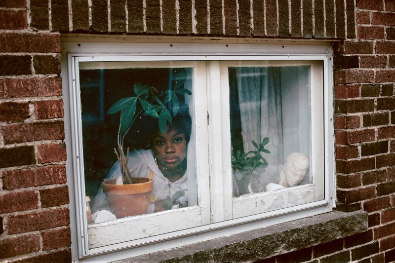 Image of a woman in a white top peering through a small window from behind a potted plant