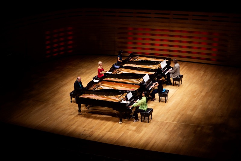 Six people are seated in front of six contiguous pianos. 