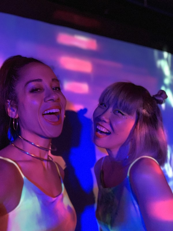 Two individuals wearing tank tops stare into the camera lens, smiling together. Projected onto their bodies are multicoloured lights. These individuals are wearing matching red lipstick and tied-up hairstyles.