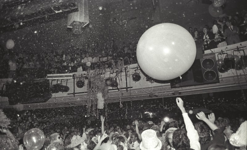 A large room. There is a crowd standing below with a series of onlooker perched upon the second floor mezzanine. A very large white ball can been floating in the center of the frame.