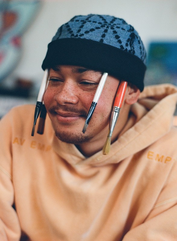 image of artist Moya wearing a pale orange hoodie and blur patterned beanie with paint brushes tucked into the hat