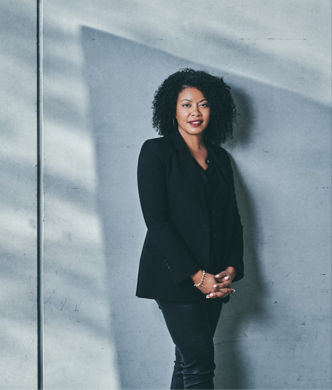 Curator Adrienne Edward wearing black jacket and trousers standing against a light blue-gray wall with diagonal lines and shadows