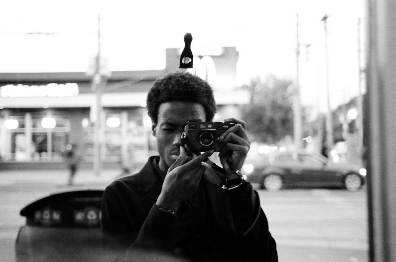 a black and white self portrait of the artist's reflection in a window, camera held to his face 