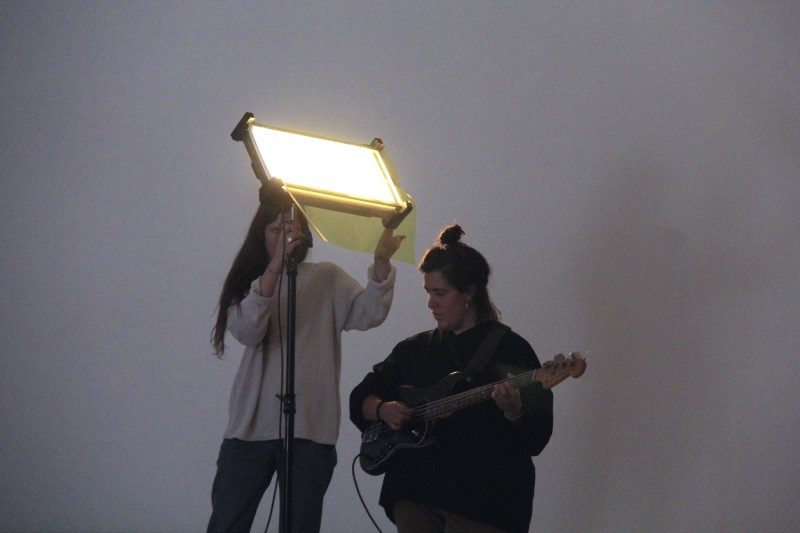 Artist Anni Spadafora standing against a grey background playing a guitar and a person standing behind her holding a light box