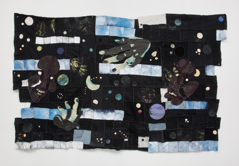 An image of a textile work by Tau Lewis, featuring recycled fabrics and leather, acrylic paint, stones, sea shell in mostly blacks, blues and whites with figures of faces and hands, 
