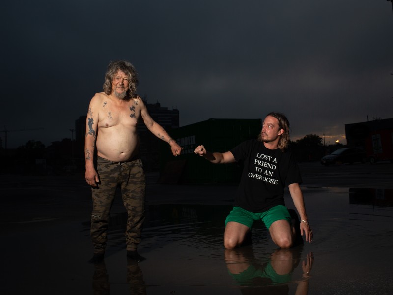 image of two men, brightly lit against a dark outdoor background. The man on the left wears camo-print pants, on the right a man wears a tshirt that reads "lost a friend to an overdose", green shorts and is kneeling. They are reaching out fists to each other. 