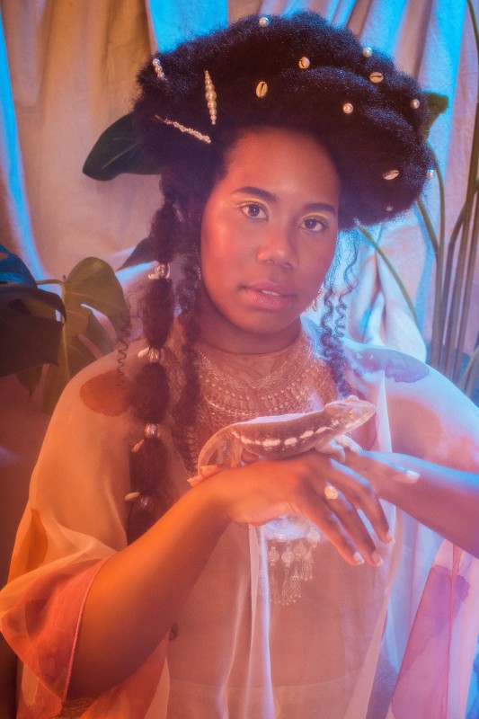 photo of DJ Gayance, with a soft focus, peach and blue textile background, wearing an etheral white gown, holding a lizard, pearls and shells dotting her hair
