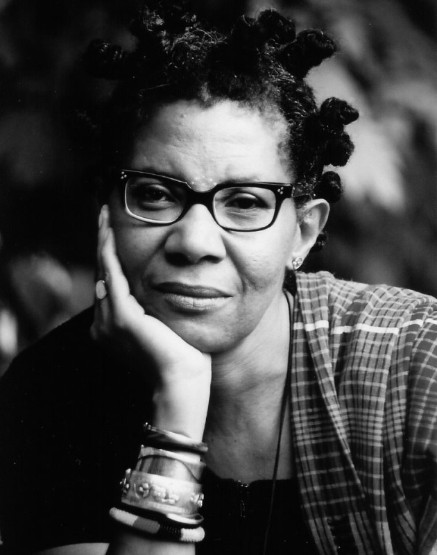 Black and white headshot of M. NourbeSe Philip wearing glasses, a striped top, holding her chin with her hand, a stack of bracelets on her wrist