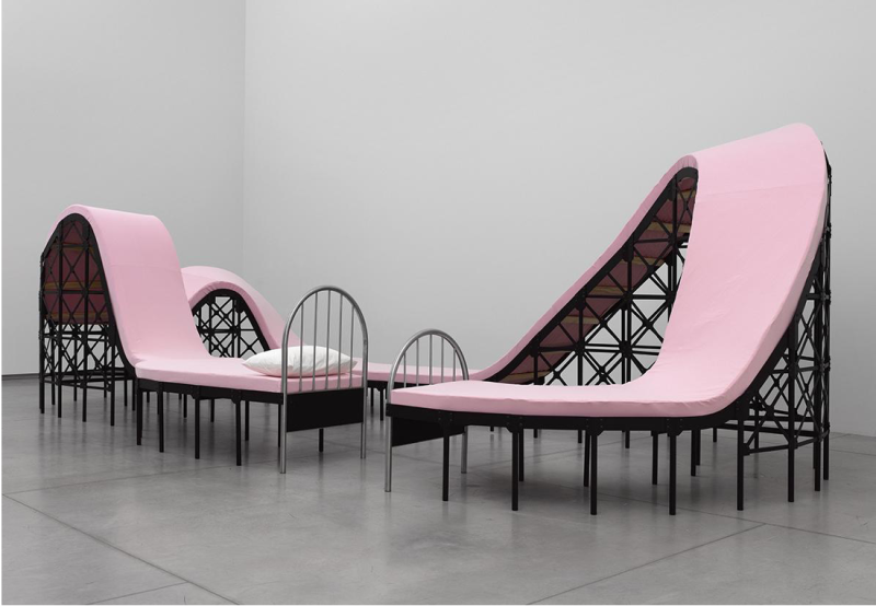 A contemporary installation of a pink bed which has been absurdly stretched, elongated and contorted into a sculpture. 