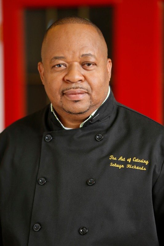 Chef Selwyn Richards wearing a black chef's uniform, looking at the viewer, with a blurred red and black background behind. 