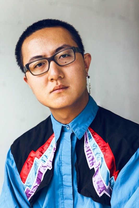 Fan Wu wearing a long earing, glasses, and a blue shirt with dark panels at the shoulder embellished with red blue and purple satin stripes