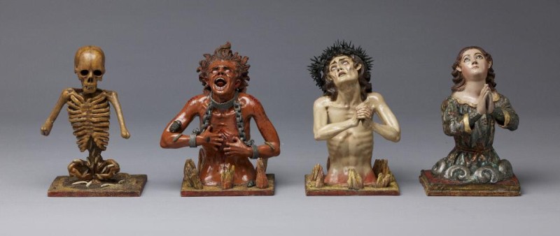 Four painted, wooden sculptures are arranged in a straight line. They are four human figures shown from the waist up. At left is a skeleton, next is a red devilish figure in chains and flames, second is a bare-chested man with fingers clasped in a begging pose, and finally, an angelic figure, in sumptuous clothing, with hands together in a prayer pose.